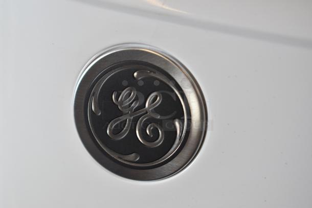 GE logo emblem on a white appliance surface, featuring metallic finish and engraved detailing, suggesting brand authenticity.