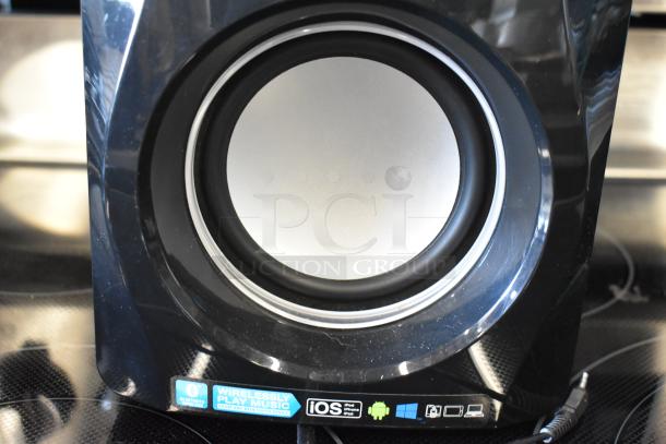 Black wireless Bluetooth speaker, circular design, compatible with iOS, Android, Windows; label indicates music streaming capability.