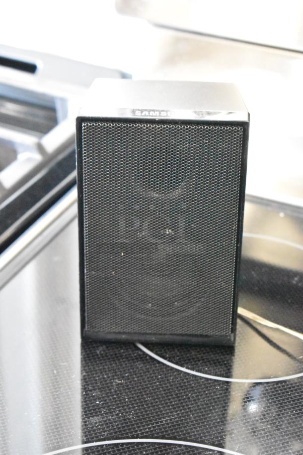 Black Samsung bookshelf speaker in good condition, metal grille front, compact design. Marked "Samsung" on top edge.
