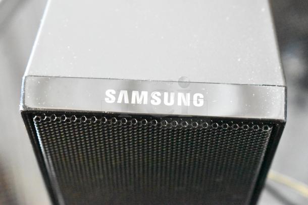 Samsung speaker, black, featuring fine metal grille. Visible dust, model unknown, prominent brand logo on the top.