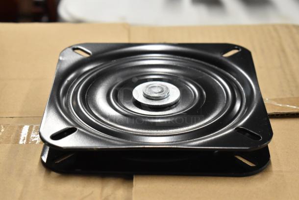 Black metal swivel plate, circular design, in new condition, with a central metal bolt, placed on a cardboard box.