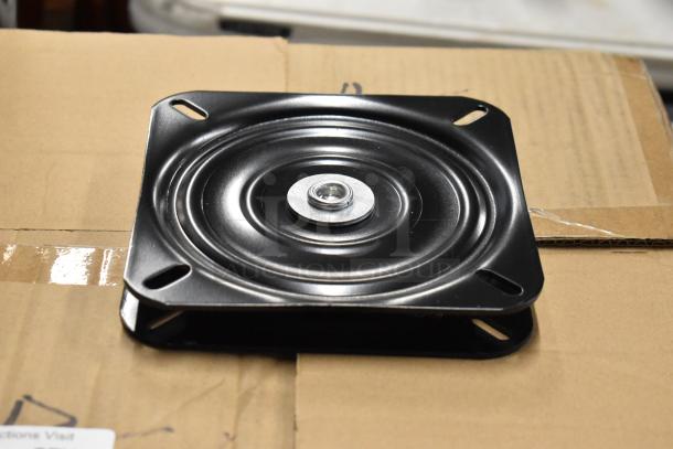 Black metal swivel plate, new condition, with bolt in center and slotted corners, on a cardboard surface.