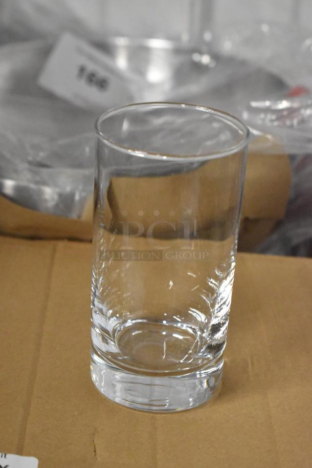 Clear glass tumbler, cylindrical shape, pristine condition, placed on a cardboard surface. Background shows blurred auction tag "166".