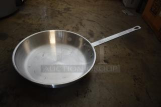 Large stainless steel frying pan with long handle and riveted attachment, showing minor scuffs, on a concrete surface.