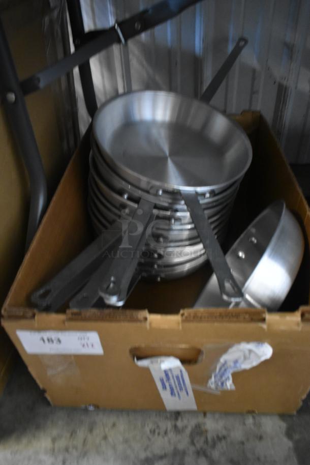 Stack of stainless steel frying pans with long handles in a cardboard box. Box has labels with unclear markings.