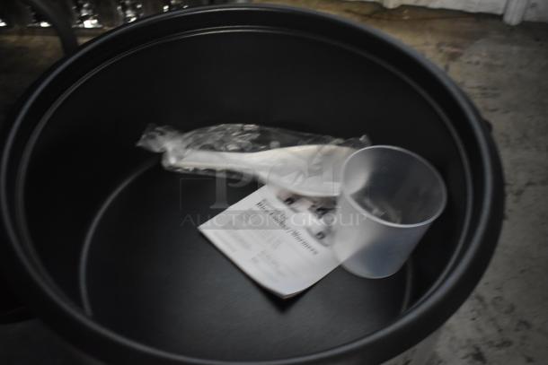 Black kitchen appliance pot with manual, plastic measuring cup, and sealed utensil inside. Appears unused, clean condition.