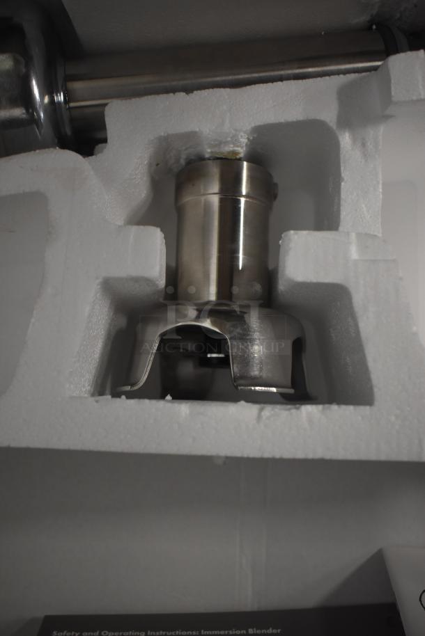 Stainless steel immersion blender base in protective packaging, appears unused. Includes operating instructions.