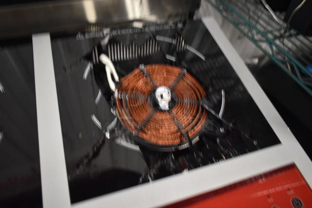 Blurred image of an induction cooktop with copper coil visible, black glass surface, and white markings. Condition unclear.