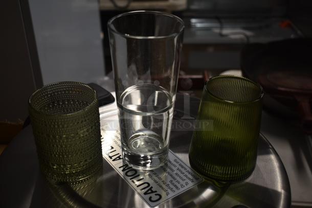 Three decorative drinking glasses, including a clear tall glass and two green textured glasses, set on a caution-labeled surface.