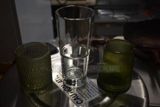 Three decorative drinking glasses, including a clear tall glass and two green textured glasses, set on a caution-labeled surface.