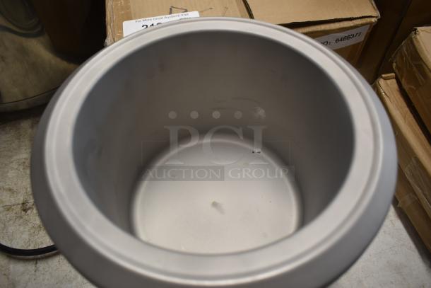 Metallic open-top container, cylindrical shape, light surface marks, placed on a concrete floor; auction label visible.