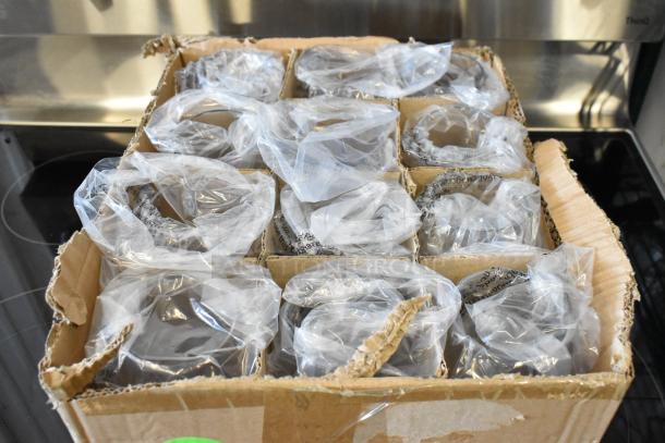 Box of glass items, each wrapped in clear plastic, arranged in a grid within a cardboard box. Items appear new.