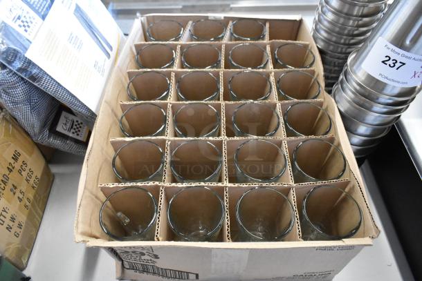 Box of 24 glass cups, neatly separated in a cardboard grid, appearing unused. Box labeled "fragile glassware" for protection.