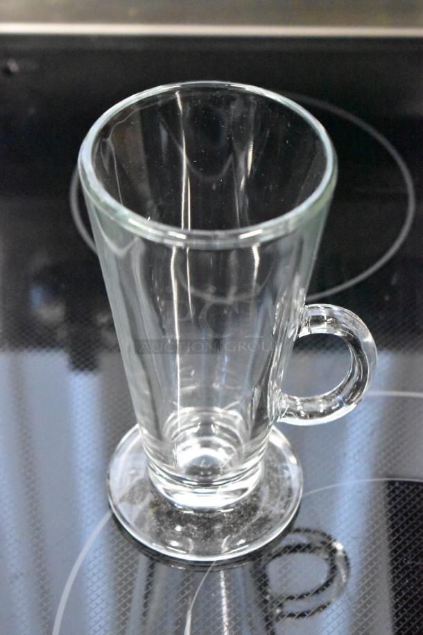 Clear glass mug with handle and flared rim, in excellent condition, displayed on a stovetop. Ideal for hot beverages.