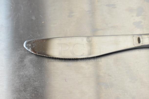 Stainless steel butter knife on metal surface, slight wear visible, sharp edge and smooth handle, no markings.