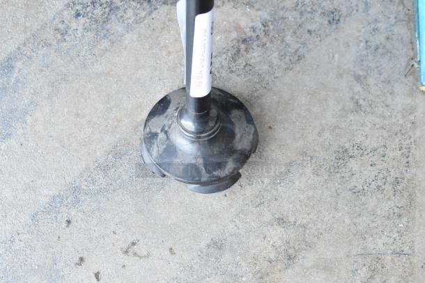 Black rubber plunger with a metal handle on a concrete floor. The handle has a label with text.