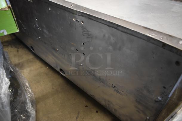 Large metal industrial box, gray with multiple small holes and signs of wear, resting on a concrete floor.