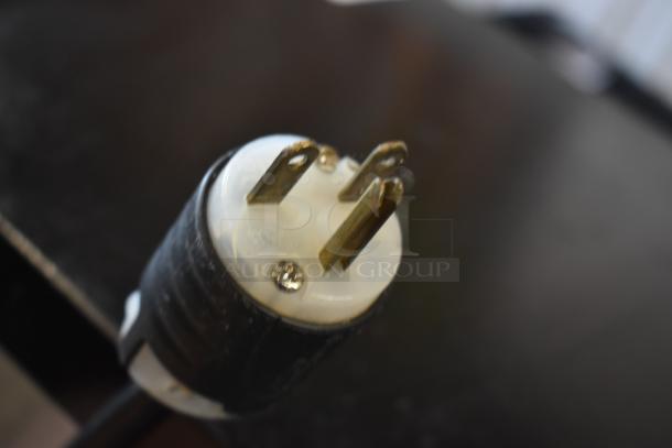 Close-up of a three-prong electrical plug with a black and white casing. The plug is in good condition, showing metal prongs and a screw fastening.