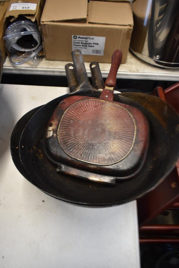 Stack of vintage cast iron skillets with visible rust, wear. Includes square skillet with red handle and circular pans.