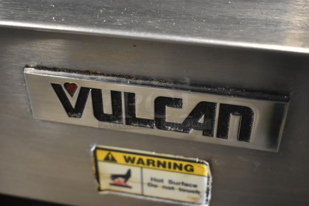 Vulcan appliance nameplate and warning label indicating hot surface. Metal finish shows slight wear and rust along edges.