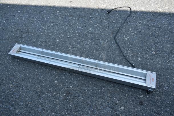 Long metal industrial heat lamp with cord on asphalt, marked "WARNING" and "Shat-R-Shield" on the end. Good condition.