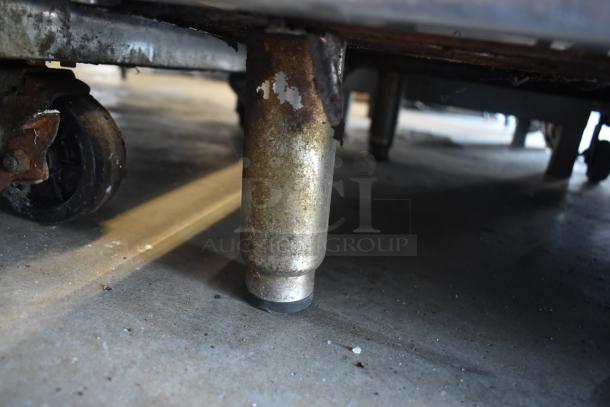 Close-up of a rusted metal caster leg on a wheeled cart, showing signs of wear. Industrial setting, concrete floor.