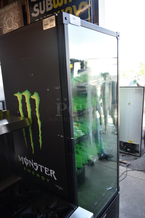 Monster Energy branded glass-door refrigerator, black exterior, used condition, with visible shelving and minor surface marks.
