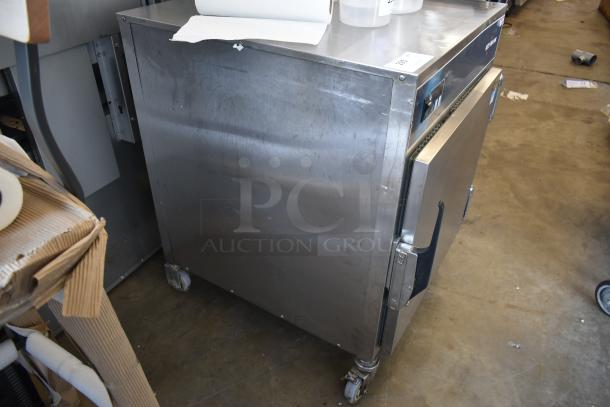 Stainless steel warming cabinet on wheels, surface wear visible, featuring a front door handle and control panel.