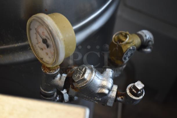 Industrial pressure gauge with metal fittings; aged condition, showing rust and wear, attached to a large tank.