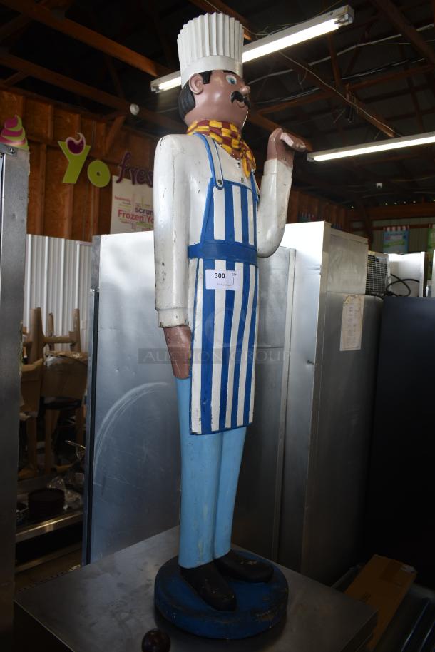 Wooden chef statue with white apron and blue stripes, chef's hat, and mustache. Display tag reads "300." Some wear visible.