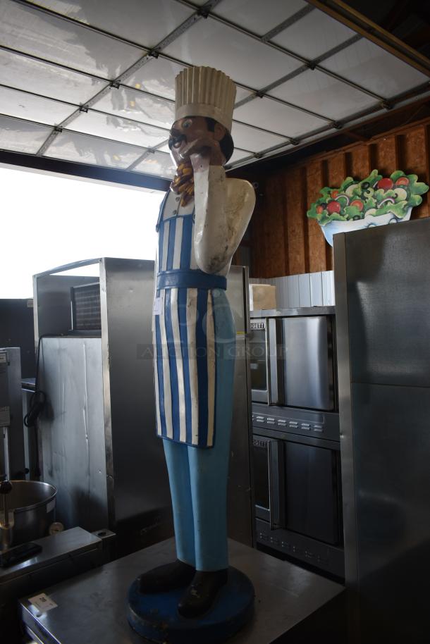 Life-size chef statue wearing a blue and white striped apron, holding a spoon, on a round base. Some wear visible.