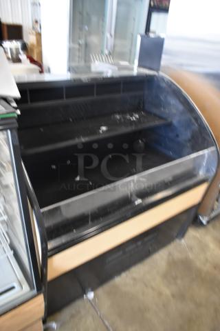 Black curved display case, two shelves, visible wear and scuffs, suitable for retail or food display, no visible labels.