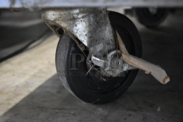 A used black caster wheel attached to a metal frame, visible dirt and wear, mounted on a concrete surface.
