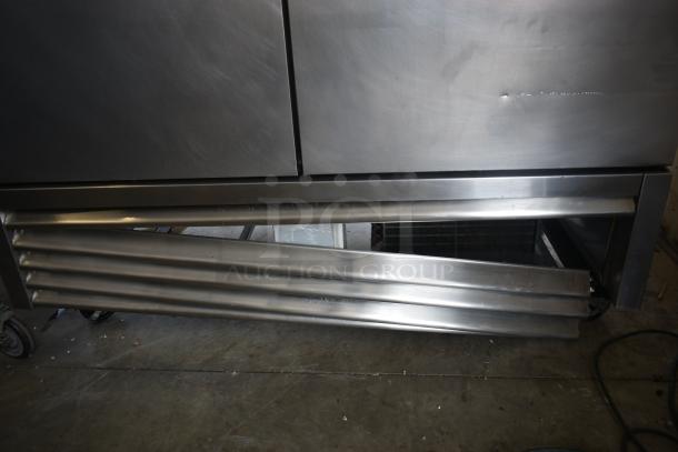 Commercial stainless steel refrigerator, visible dents and scratches, lower vent panel partially missing, on wheels.