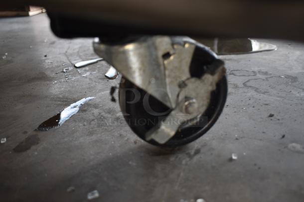Close-up of a metal caster wheel on a concrete floor, showing signs of use and some rust. Surface appears slightly wet.