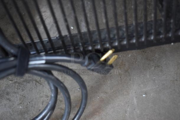 Coiled black power cord with a two-prong plug, resting on a concrete floor near a metal grate.