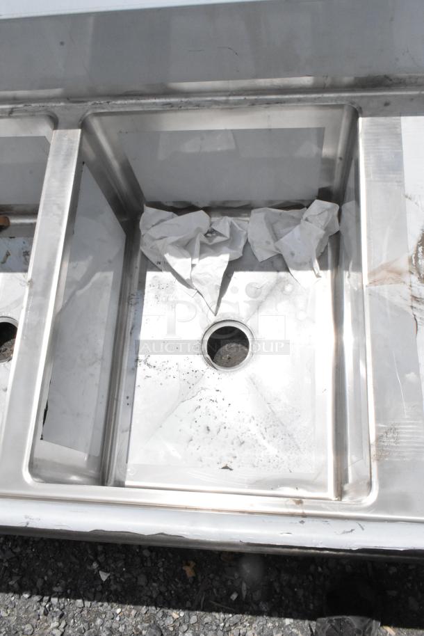 Stainless steel triple sink with drains, slight surface wear, minor dirt and paper debris present. Ideal for commercial use.