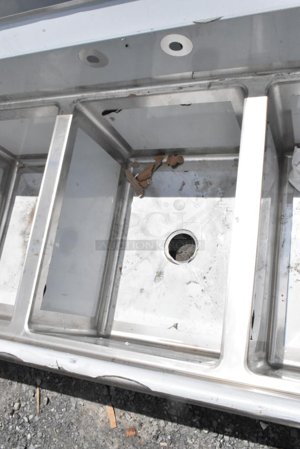 Stainless steel industrial sink with multiple basins, visible wear, and debris. Drain holes and metal frame visible.