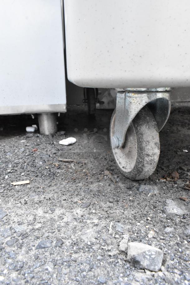 Close-up of metal caster wheel attached to a large, white industrial cart. Wheel shows signs of wear, set on rough concrete ground.