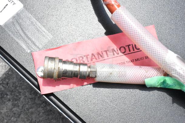Metal hose fitting on textured surface, attached to white braided hose. Includes red "IMPORTANT NOTICE" tag.