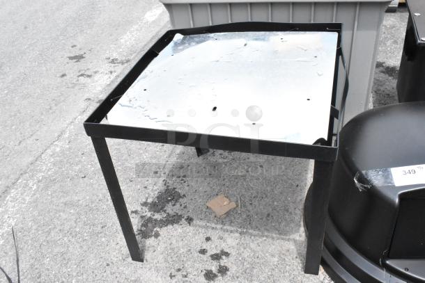 Black metal table with a reflective top, in used condition, showing some surface wear and dents, placed outdoors.