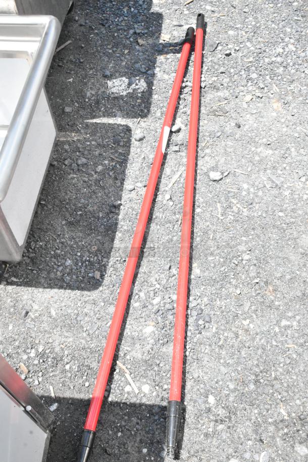 Two red extension poles with black handles, lying on gravel. Moderate wear, one with a white label.