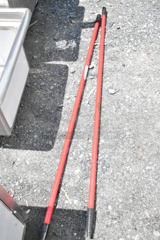 Two red extension poles with black handles, lying on gravel. Moderate wear, one with a white label.