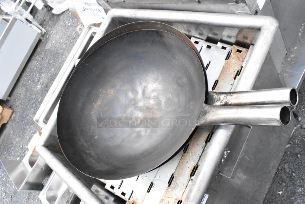 Large industrial double-handled wok, shows signs of wear and rust, placed on a metal rack at an auction site.