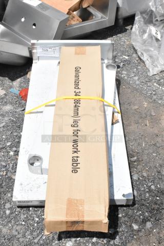 Cardboard box labeled "Galvanized 34-inch leg for work table" with metal brackets on gravel. Item tag present.
