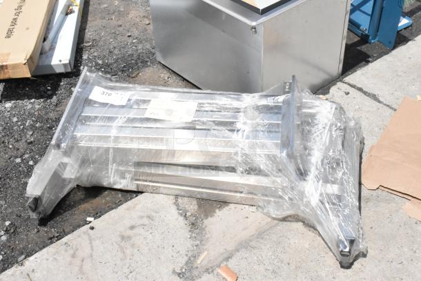 Stacked metal shelving unit wrapped in plastic with labels indicating model numbers, positioned on outdoor pavement.