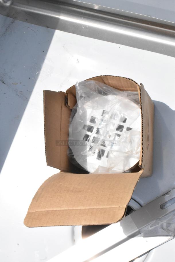 Open box containing a new metal ventilation grille in plastic wrapping, resting on a reflective surface.