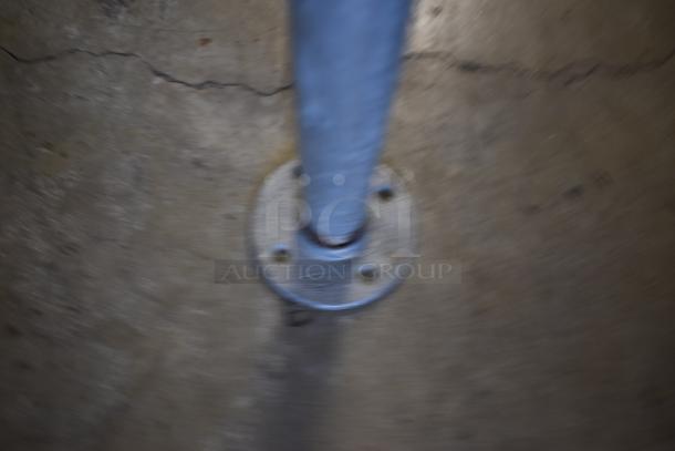 Blurry image of a metallic pole with a base plate on a concrete surface. No visible markings or labels.