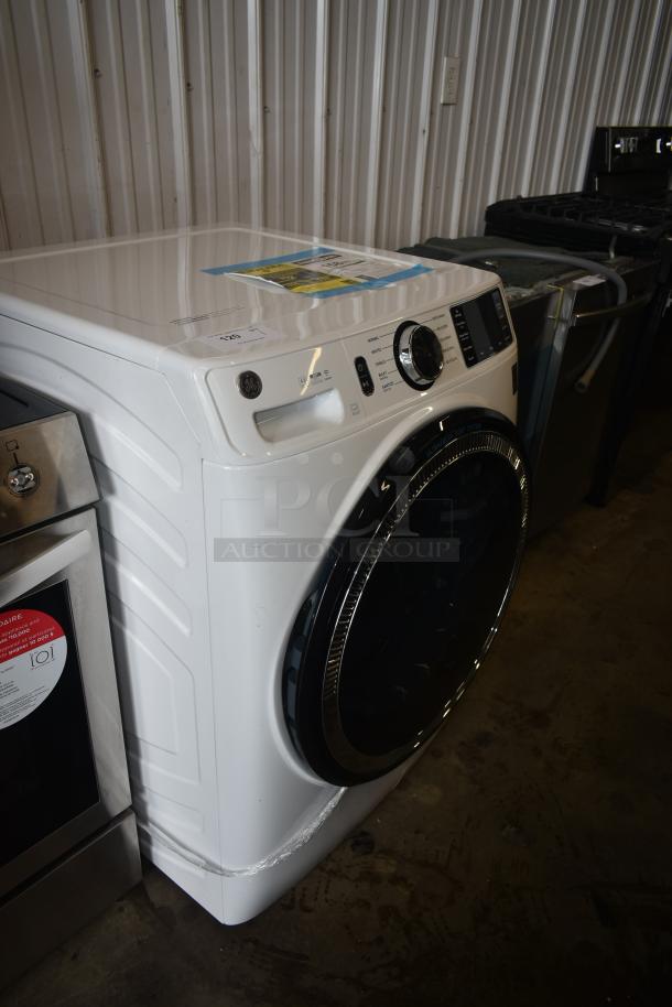 White GE front-load washer with large black door and digital controls. Model on display in auction with minor scuff marks.