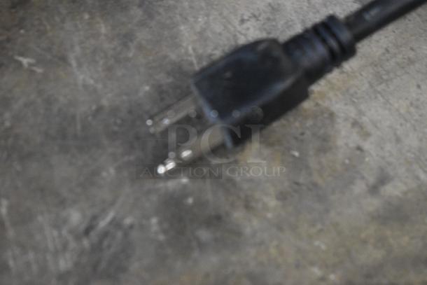 Blurry image of a black electrical plug with two metal prongs on a rough surface. Condition and other details unclear.